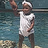 child, toddler, baby, outdoor, poolside, water, swimming_pool, white_outfit, dress, headband, bow, sandals, standing, holding_hand, adult_hand, portrait, sunlight, dock, rocks, happy