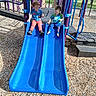 children, toddler, playground, slide, outdoor, park, siblings, sitting, play_structure, blue_slide, sunlight, wood_chips, summer, clothing, smiling, childhood, recreation, playtime,  안전한, photography