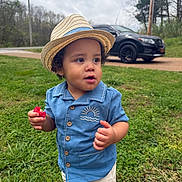 Ashton is registered to the contest to win money with this photo: toddler, child, hat, straw_hat, blue_shirt, shorts, grass, outdoor, daytime, vehicle, black_suv, food, holding, nature, trees, cloudy_sky, road, person, cute, summer_clothing