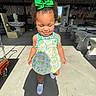 child, toddler, smiling, green_bow, lemon_dress, white_shoes, shadow, sunlight, outdoor, stone_sculptures, workshop, garden_furniture, concrete_floor, curly_hair, closed_eyes, earrings, happy, portrait, walking, playful