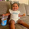 toddler, child, smiling, sitting, couch, headband, snack, container, spoon, shirt, text_on_clothing, indoor, cozy, happy, person, baby, arm, leg, foot, home