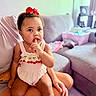 adult_hand, baby, basket, child, couch, curly_hair, cute, doll, finger_in_mouth, home, indoor, lap, living_room, person, pink_clothing, red_bow, sitting, sofa, toddler, toy