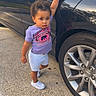 black_car, car, casual_clothing, child, concrete, curly_hair, daylight, door_handle, earrings, outdoor, person, purple_shirt, reflection, shorts, sidewalk, standing, tire, toddler, wheel, white_shoes