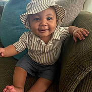 Hakeem is registered to the contest to win money with this photo: baby, child, smiling, hat, striped_clothing, shorts, couch, pillows, indoor, happy, portrait, sitting, skin, face, hand, foot, home, cute, infant, cozy