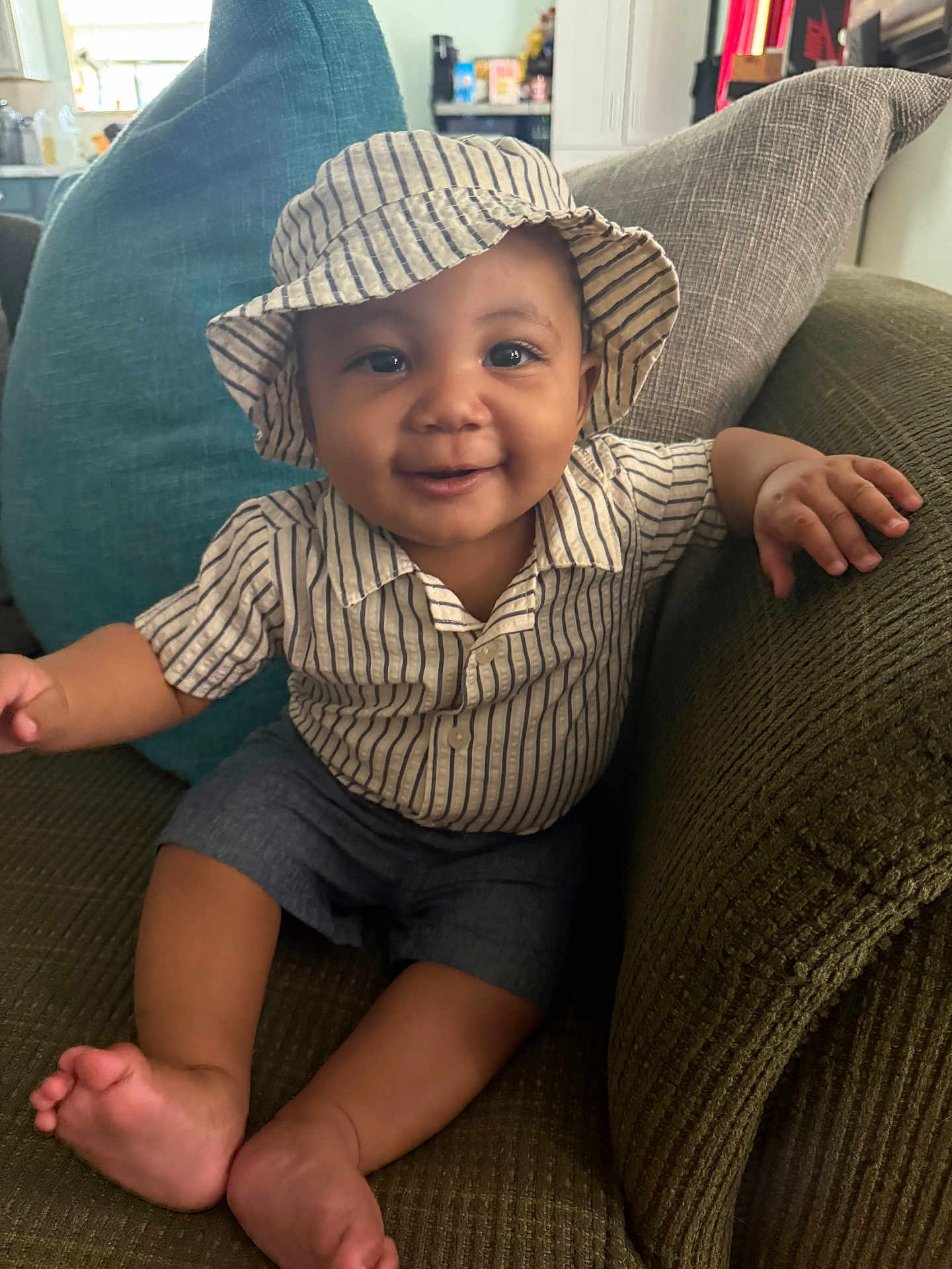 Hakeem is registered to the contest to win money with this photo: baby, child, smiling, hat, striped_clothing, shorts, couch, pillows, indoor, happy, portrait, sitting, skin, face, hand, foot, home, cute, infant, cozy