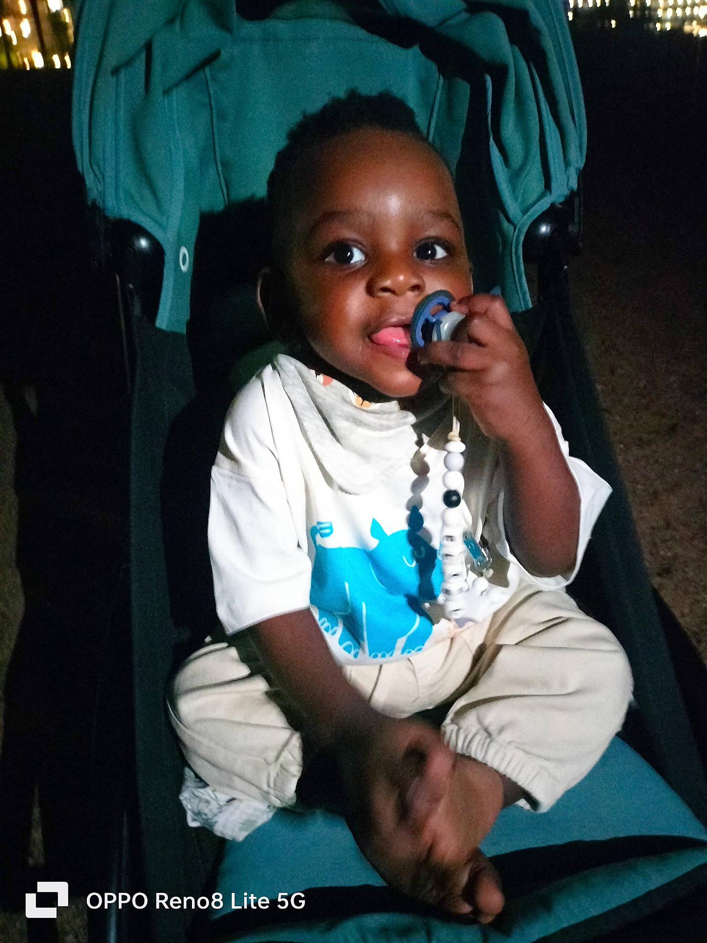 éliel participe au concours pour gagner de l'argent avec cette photo : baby, barefoot, beads, beige_pants, blue_print, child, cute, dark_background, indoors, infant, night, pacifier, playful, portrait, sitting, smiling, stroller, toddler, white_shirt, young_child