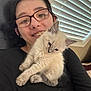 person, glasses, smile, kitten, cat, indoor, window_blinds, black_shirt, fur, pet, cute, young_adult, face, hair, cozy, holding, resting, light, comfort, feline