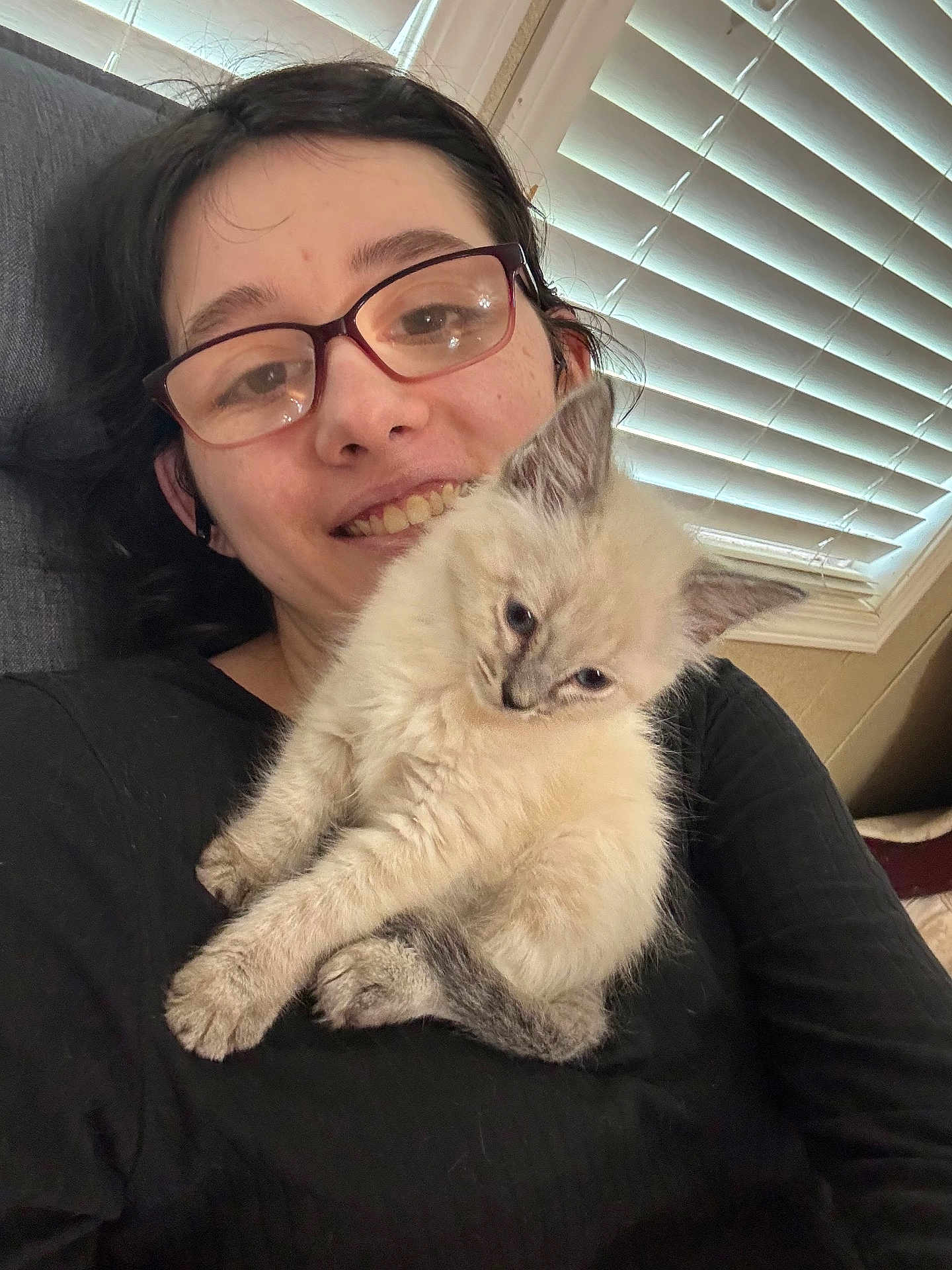 Zoey is registered to the contest to win money with this photo: person, glasses, smile, kitten, cat, indoor, window_blinds, black_shirt, fur, pet, cute, young_adult, face, hair, cozy, holding, resting, light, comfort, feline
