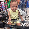 aisle, baby, black_strap, blanket, child, drink_cans, expression, face, footwear_absent, grocery_store, hand, indoor, person, retail, shelf, shopping_cart, sitting, supermarket, toddler, yellow_clothing