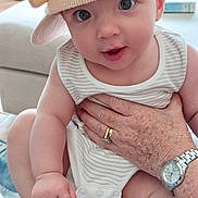Emilia joined the competition — help win amazing prizes! baby, infant, hat, striped_clothing, onesie, hand, adult_hand, watch, ring, freckles, indoor, couch, person, face, eyes, legs, foot, expression, cute, child
