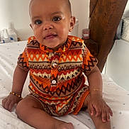 Jelany a rejoint le concours — aidez-le/la à gagner de superbes lots ! baby, child, infant, sitting, bed, clothing, orange, patterned_clothes, bracelet, wooden_beam, indoors, smiling, face, cute, person, home, decor, foot, hand, curious