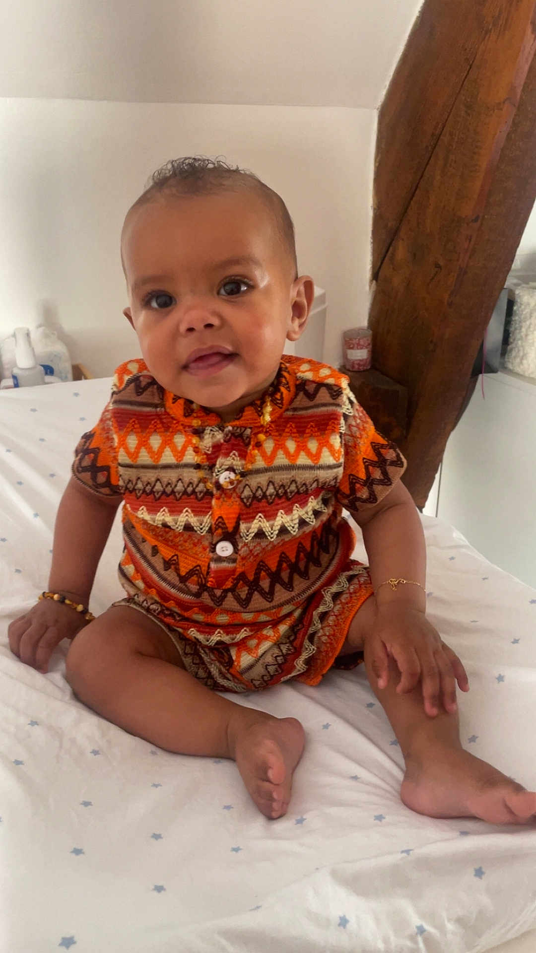 Jelany a rejoint le concours — aidez-le/la à gagner de superbes lots ! baby, child, infant, sitting, bed, clothing, orange, patterned_clothes, bracelet, wooden_beam, indoors, smiling, face, cute, person, home, decor, foot, hand, curious