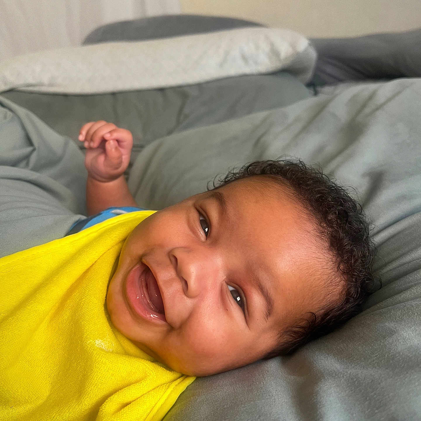 Rylo is registered to the contest to win money with this photo: baby, bed, blanket, child, closeup, comfort, curly_hair, cute, expression, face, happy, indoors, infant, lying_down, person, portrait, relaxed, skin, smiling, yellow_bib