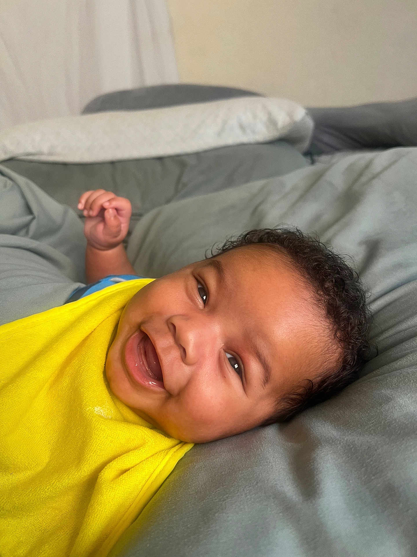 Rylo is registered to the contest to win money with this photo: baby, smiling, yellow_bib, curly_hair, face, happy, infant, lying_down, bed, blanket, closeup, child, skin, portrait, indoors, relaxed, cute, comfort, expression, person
