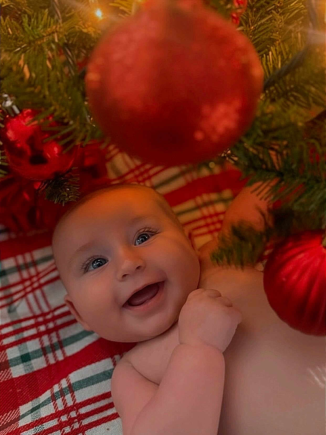 Parker is registered to the contest to win money with this photo: baby, smiling, face, christmas_tree, christmas_ornament, red_ornament, plaid_blanket, holiday, festive, infant, cute, cheerful, indoors, closeup, skin, child, celebration, decorations, happy, lying_down