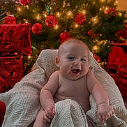 Parker is registered to the contest to win money with this photo: baby, blanket, christmas_tree, christmas_lights, red_ornaments, gifts, holiday, festive, smiling, laughing, infant, cheerful, indoors, decorations, celebration, warm, cozy, wrapped, sitting, cute