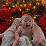 baby, blanket, christmas_tree, christmas_lights, red_ornaments, gifts, holiday, festive, smiling, laughing, infant, cheerful, indoors, decorations, celebration, warm, cozy, wrapped, sitting, cute
