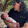 man, baby, newborn, outdoor, nature, trees, forest, holding, sleeping, person, hat, facial_hair, black_shirt, blue_shorts, rocky_ground, arm, hand, love, tenderness, calm