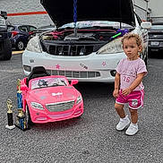 Cyri-Faith is registered to the contest to win money with this photo: toddler, child, toy_car, trophies, white_car, hood_open, parking_lot, cloudy_sky, asphalt, shorts, tshirt, sneakers, curly_hair, outdoor, vehicle, car_mechanic, small_car, award, person, daytime