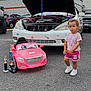toddler, child, toy_car, trophies, white_car, hood_open, parking_lot, cloudy_sky, asphalt, shorts, tshirt, sneakers, curly_hair, outdoor, vehicle, car_mechanic, small_car, award, person, daytime