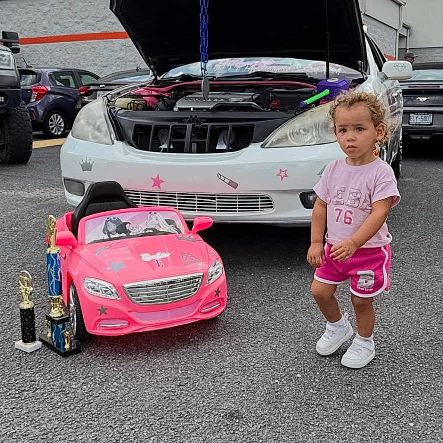 Cyri-Faith is registered to the contest to win money with this photo: asphalt, award, car_mechanic, child, cloudy_sky, curly_hair, daytime, hood_open, outdoor, parking_lot, person, shorts, small_car, sneakers, toddler, toy_car, trophies, tshirt, vehicle, white_car