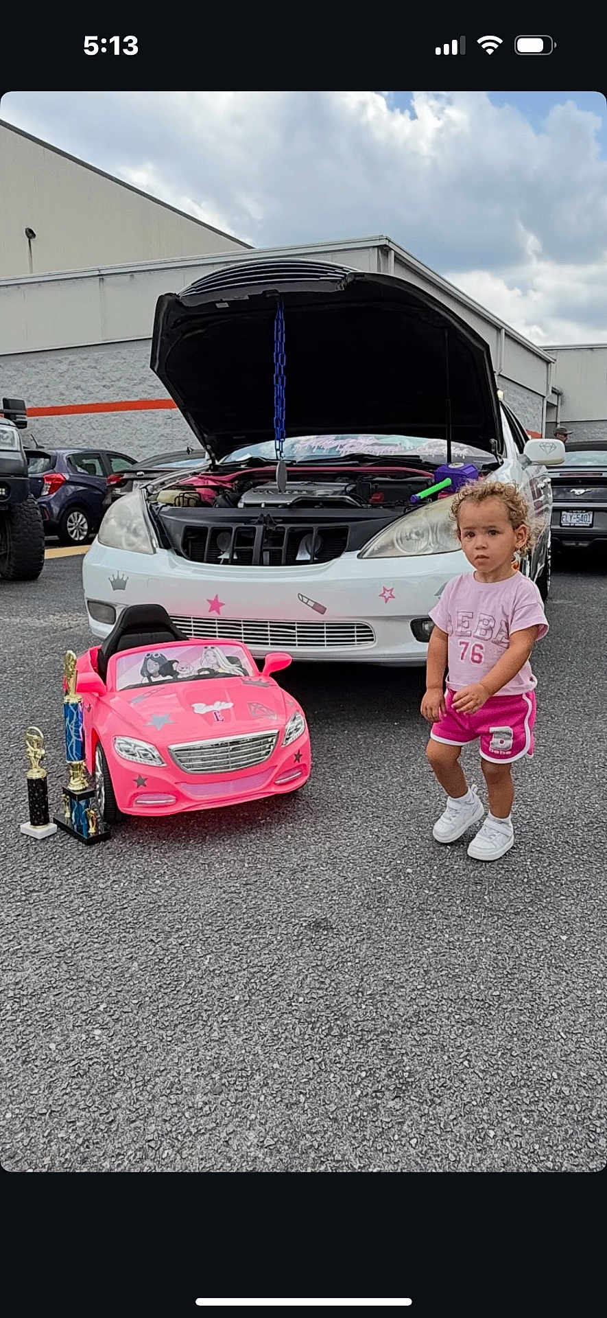 Cyri-Faith is registered to the contest to win money with this photo: toddler, child, toy_car, trophies, white_car, hood_open, parking_lot, cloudy_sky, asphalt, shorts, tshirt, sneakers, curly_hair, outdoor, vehicle, car_mechanic, small_car, award, person, daytime