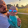 Athena joined the competition — help win amazing prizes! baby, child, man, sunglasses, tattoo, field, sunset, outdoor, grass, person, reflection, holding, casual, daylight, sky, trees, sports_field, clothing, portrait, cute
