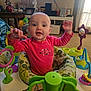 baby, child, infant, pink_shirt, activity_center, toys, smiling, arms_up, living_room, lamp, photoframes, curtains, carpet, furniture, home_interior, play, fun, happy, face, person