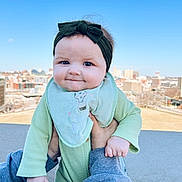 Brylee joined the competition — help win amazing prizes! adult_hands, baby, bib, blue_sky, building, cheeks, cityscape, clothing, concrete_wall, cute, daylight, green_outfit, hand, headband, infant, outdoor, portrait, sky, smile, urban