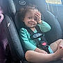 toddler, child, car_seat, curly_hair, smile, hand_on_face, seat_belt, car_interior, window, clothing, green_sweater, baby, sitting, safety, cute, indoors, daylight, happy, person, seat