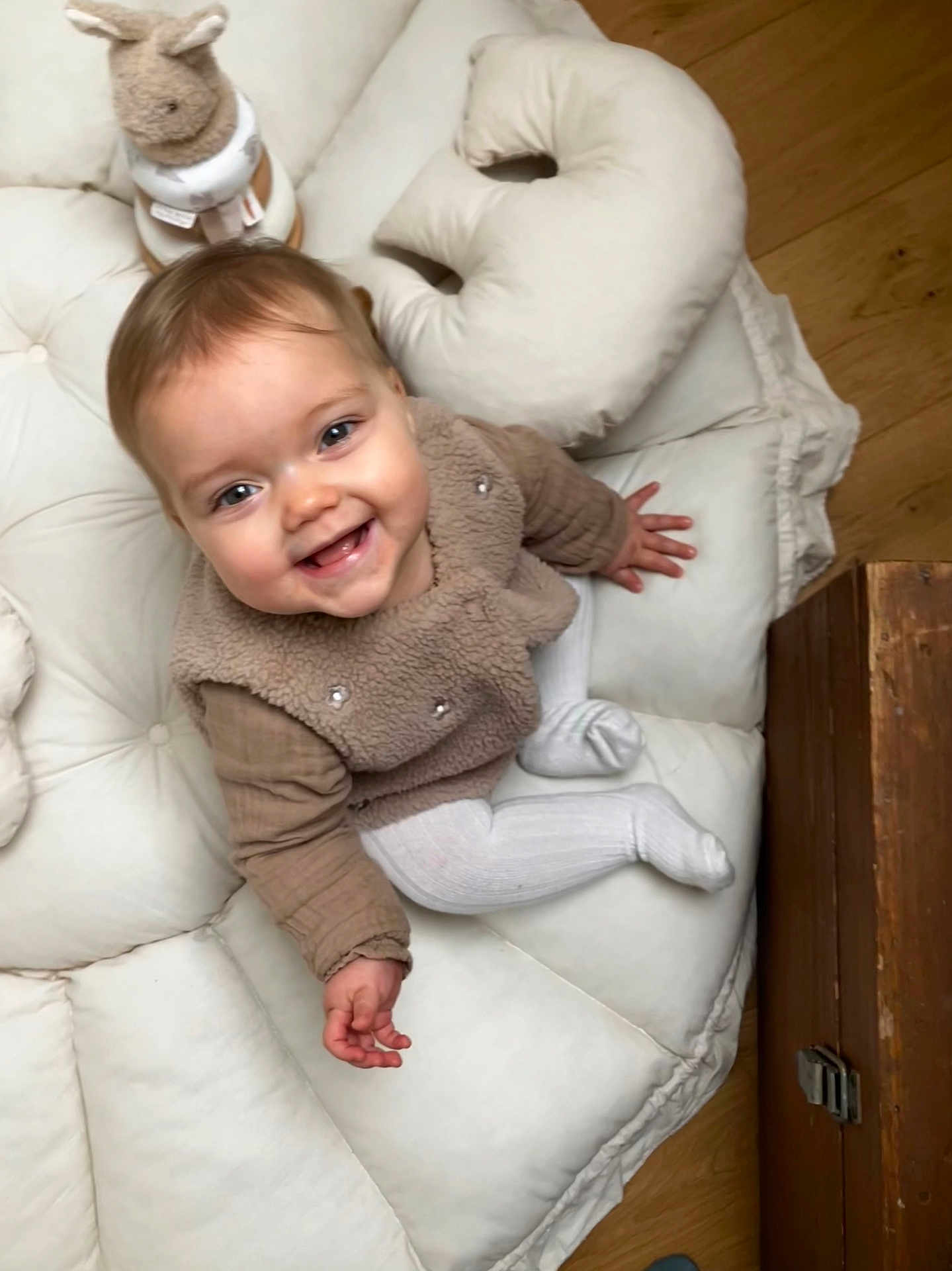 Romy participe au concours pour gagner de l'argent avec cette photo : baby, smiling, child, cushion, plush_toy, indoor, white, brown, floor, wooden_floor, happy, cute, toddler, sitting, cozy, soft, clothing, toy, face, infant