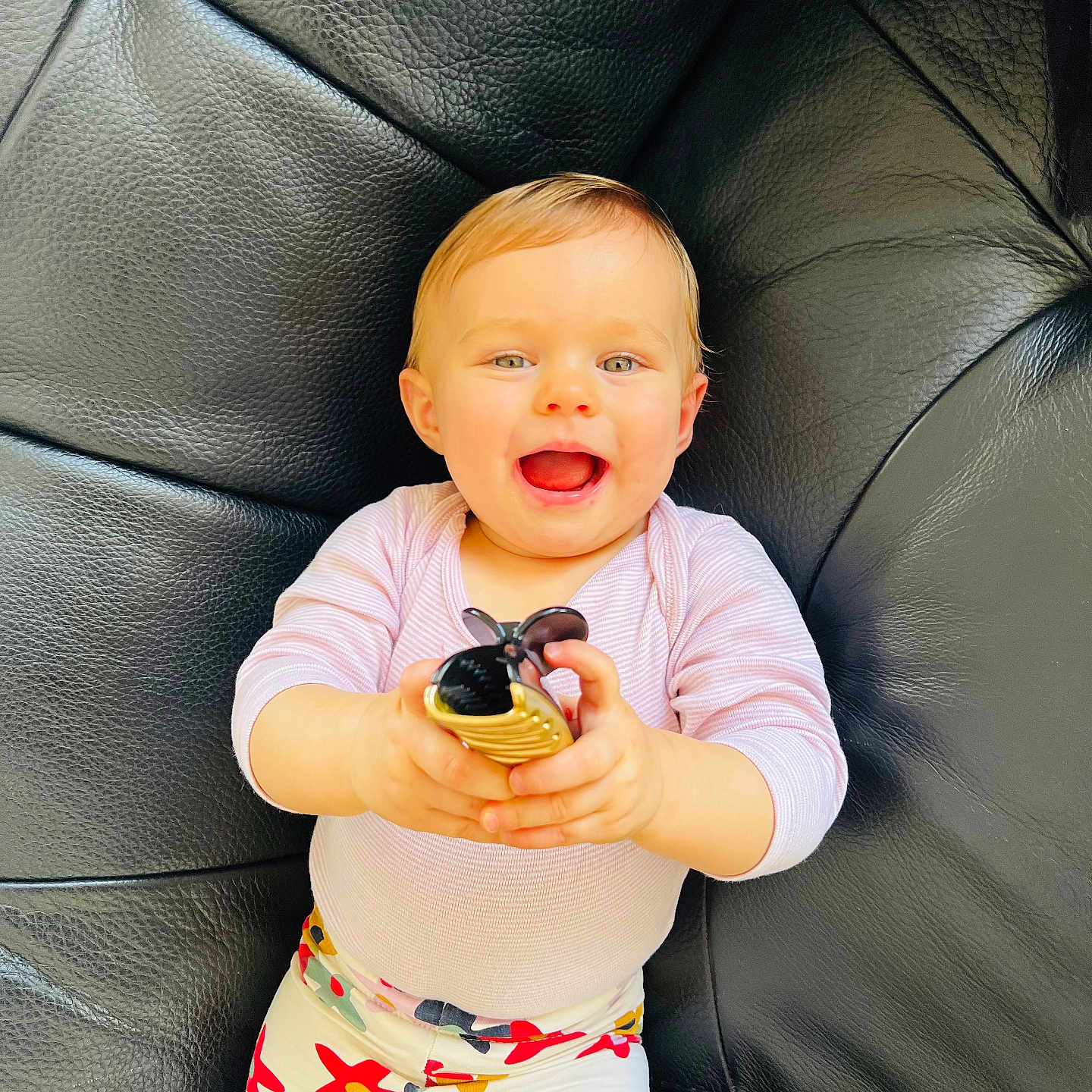 Charline participe au concours pour gagner de l'argent avec cette photo : baby, black_leather_couch, bright_eyes, casual_clothing, child, colorful_pants, comfortable, cute, floral_pattern, fun, happy, holding_object, indoor, person, playful, portrait, sitting, smiling, toy, young_child