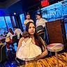 Kimberly is registered to the contest to win money with this photo: woman, baby, dress, bow, restaurant, table, people, indoor, long_hair, holding, child, furniture, light, exit_sign, casual, conversation, seated, social, background, dining