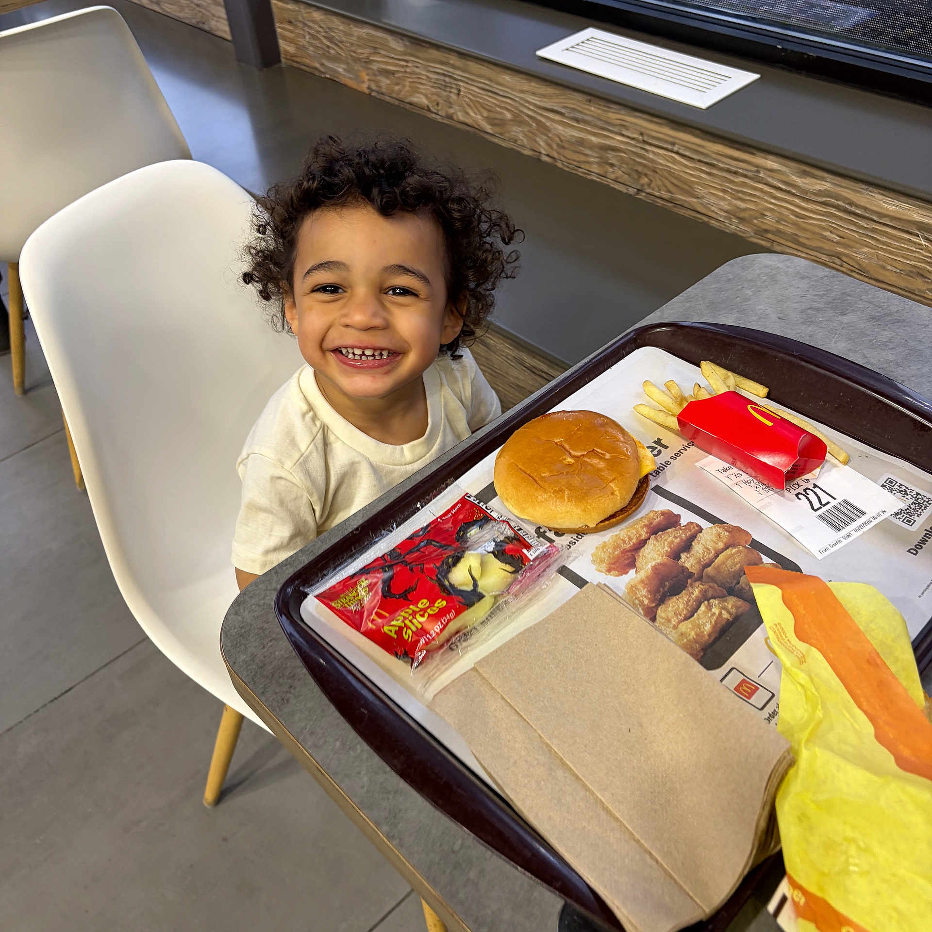 Hezekiah is registered to the contest to win money with this photo: apple_slices, burger, chair, child, curly_hair, fast_food, food_packaging, fries, happy, indoor, meal, modern_interior, napkin, person, restaurant, smiling, table, toddler, tray, window