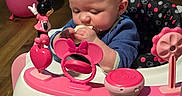 Zoey is registered to the contest to win money with this photo: activity_center, baby, baby_walker, ball, blue_clothing, chewing, child, close_up, dumbbell, eating, face, hands, hardwood_floor, indoor, infant, minnie_mouse, pink, seat, sitting, toy