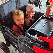 Chloe is registered to the contest to win money with this photo: toddler, child, stroller, helmet, snack, bottle, double_stroller, outdoor, people, legs, concrete_floor, clothing, seat, baby, protective_gear, red, black, pink, person, casual
