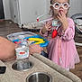 child, girl, toy, doctor, glasses, pink_dress, playing, indoor, person, dog, pet, bed, floor, hand, water_bottle, sofa, cup_holder, remote_control, wood_floor, home