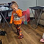 child, toddler, orange_shirt, patterned_pants, wooden_floor, living_room, floor_lamp, toy_box, television, slipper, cat, furniture, home, cute_pose, indoor, casual_clothing, floor, smiling, person, cozy