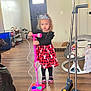 Chloe And Zoey is registered to the contest to win money with this photo: child, girl, tiara, toy_microphone, dress, black_top, indoor, wood_floor, broom, dustpan, baby_swing, footwear, curious, standing, living_room, household_items, casual, home, floor, playtime