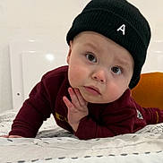 Anthuan is registered to the contest to win money with this photo: baby, child, face, beanie, hat, clothing, maroon, bed, blanket, hand, portrait, indoor, expression, curious, looking, person, young_child, cozy, casual, resting
