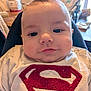 apparel, baby, chubby_cheeks, closeup, clothing, cute, domestic, expression, face, furniture, high_chair, indoor, infant, person, portrait, sitting, soft_lighting, superman_logo, table, young_child