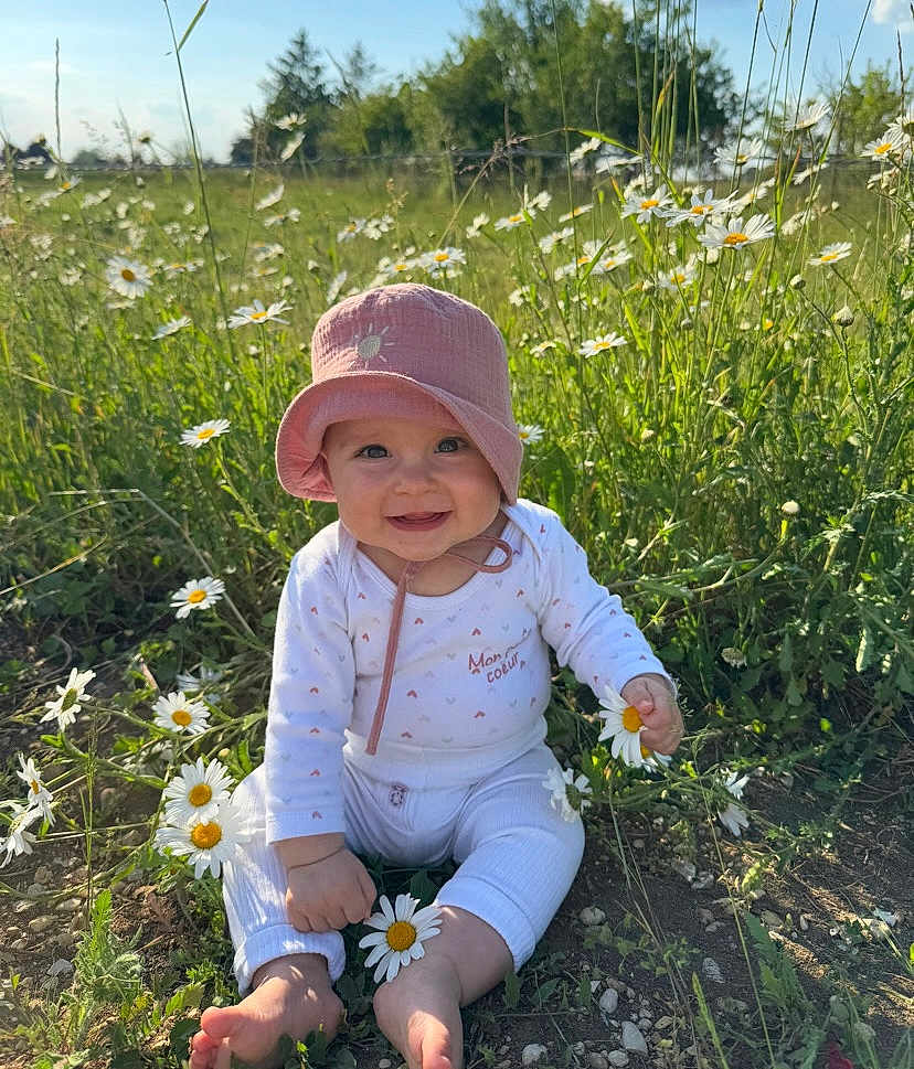 Romy a rejoint le concours — aidez-le/la à gagner de superbes lots ! baby, child, pink_hat, daisies, flowers, grass, outdoor, nature, sunlight, greenery, barefoot, smiling, happy, sitting, field, spring, casual_clothing, cute, infant, playful