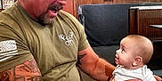 Miles is registered to the contest to win money with this photo: baby, beard, cap, casual, curiosity, food, indoor, infant, interaction, man, person, plate, restaurant, seated, short_sleeve_shirt, smile, sports_memorabilia, table, tattoo, wood_paneling