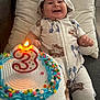 baby, smiling, hoodie, birthday_cake, candle, number_3, celebration, confetti, colorful, happy, infant, sitting, pillow, blanket, indoors, cute, food, party, child, fun