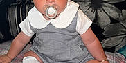 Beau-Harley is registered to the contest to win money with this photo: baby, child, pacifier, blue_eyes, socks, plaid_blanket, gray_outfit, white_collar, sitting, indoor, cushion, pillow, floral_pattern, striped_cushion, curly_hair, cute, portrait, person, infant, cozy