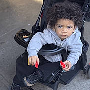 Jordan joined the competition — help win amazing prizes! child, toddler, stroller, curly_hair, hoodie, black_pants, sneakers, snack, red_packet, outdoor, sidewalk, serious_expression, person, baby, seat, wheel, jacket, holding, young_child, urban