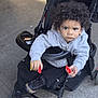 child, toddler, stroller, curly_hair, hoodie, black_pants, sneakers, snack, red_packet, outdoor, sidewalk, serious_expression, person, baby, seat, wheel, jacket, holding, young_child, urban