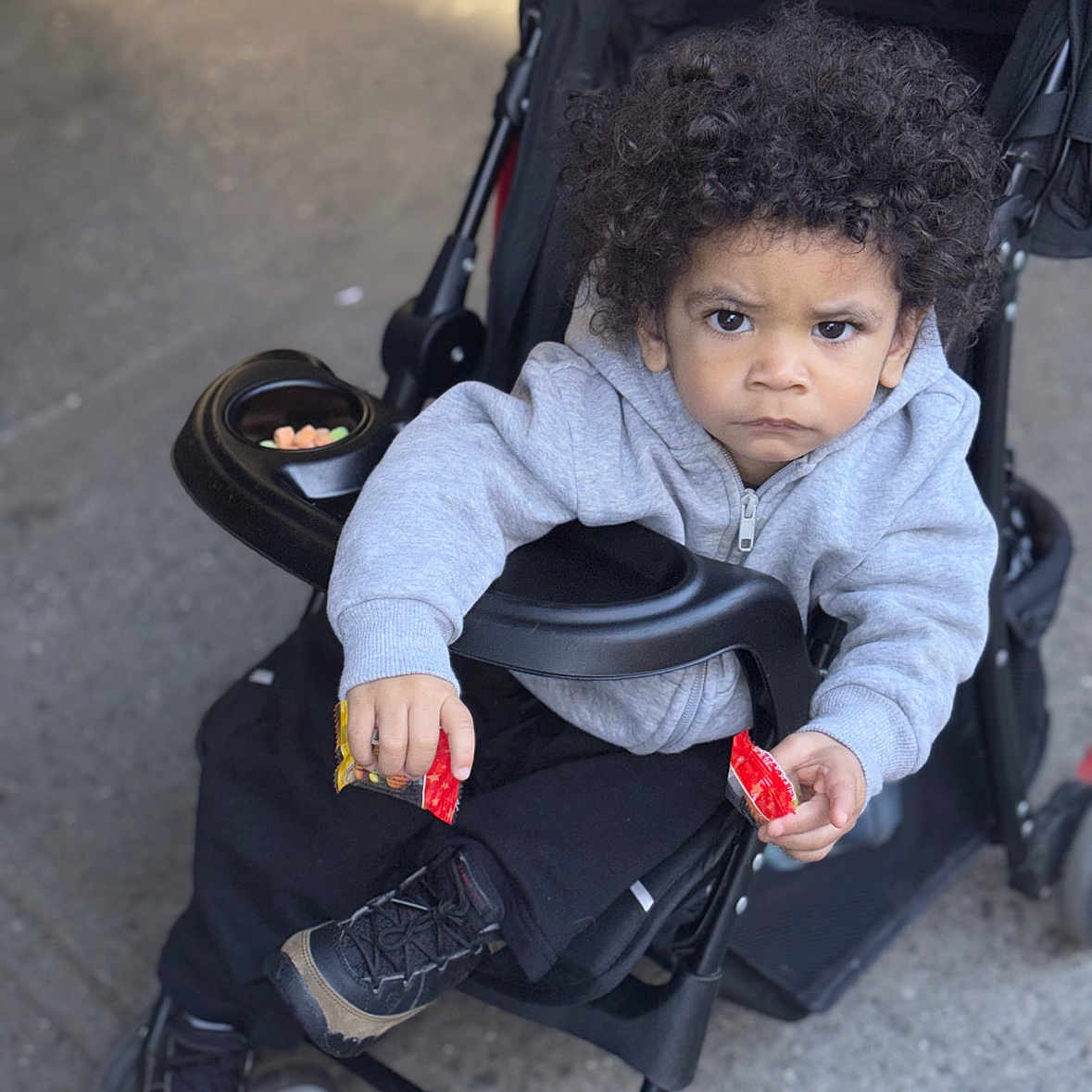 Jordan joined the competition — help win amazing prizes! baby, black_pants, child, curly_hair, holding, hoodie, jacket, outdoor, person, red_packet, seat, serious_expression, sidewalk, snack, sneakers, stroller, toddler, urban, wheel, young_child