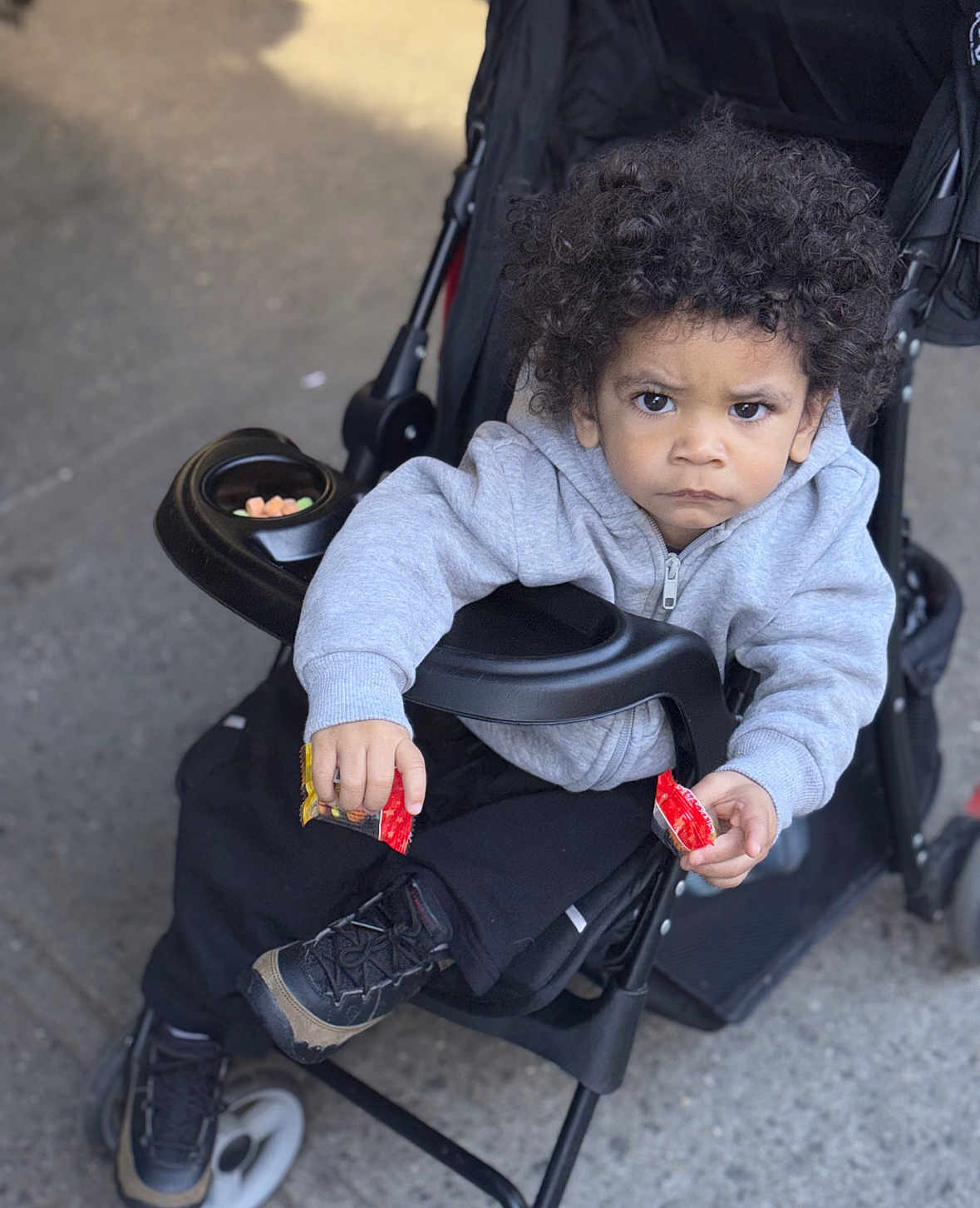 Jordan joined the competition — help win amazing prizes! child, toddler, stroller, curly_hair, hoodie, black_pants, sneakers, snack, red_packet, outdoor, sidewalk, serious_expression, person, baby, seat, wheel, jacket, holding, young_child, urban