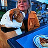 affection, background, blue_sky, bulldog, casual, chair, daylight, dog, food, hand, orange_jacket, outdoor, person, pet, pie, plate, seating_area, table, woman, wooden_shed